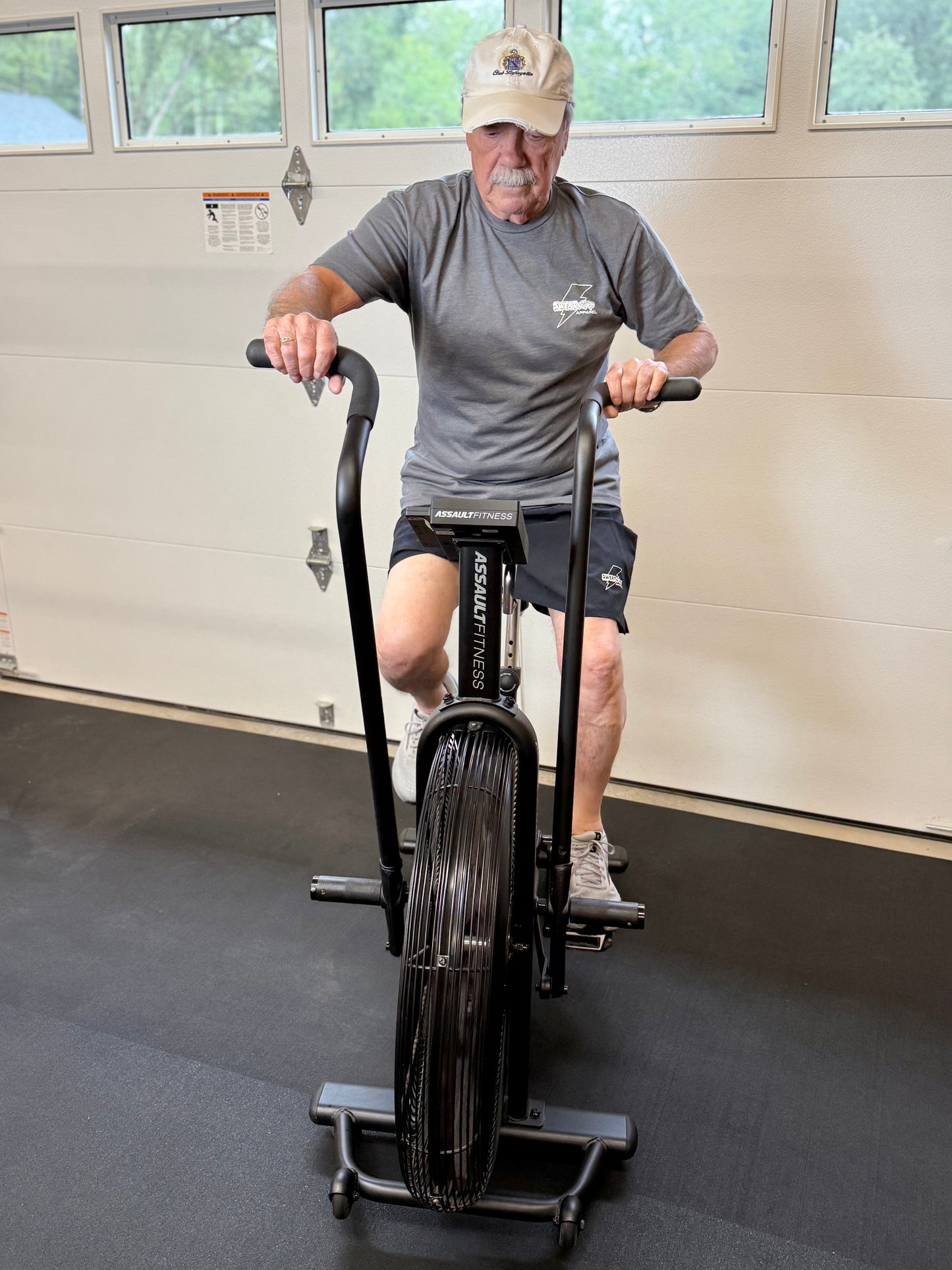 Man exercising on a stationary bike indoors wearing gray Sweat Storm Apparel t-shirt