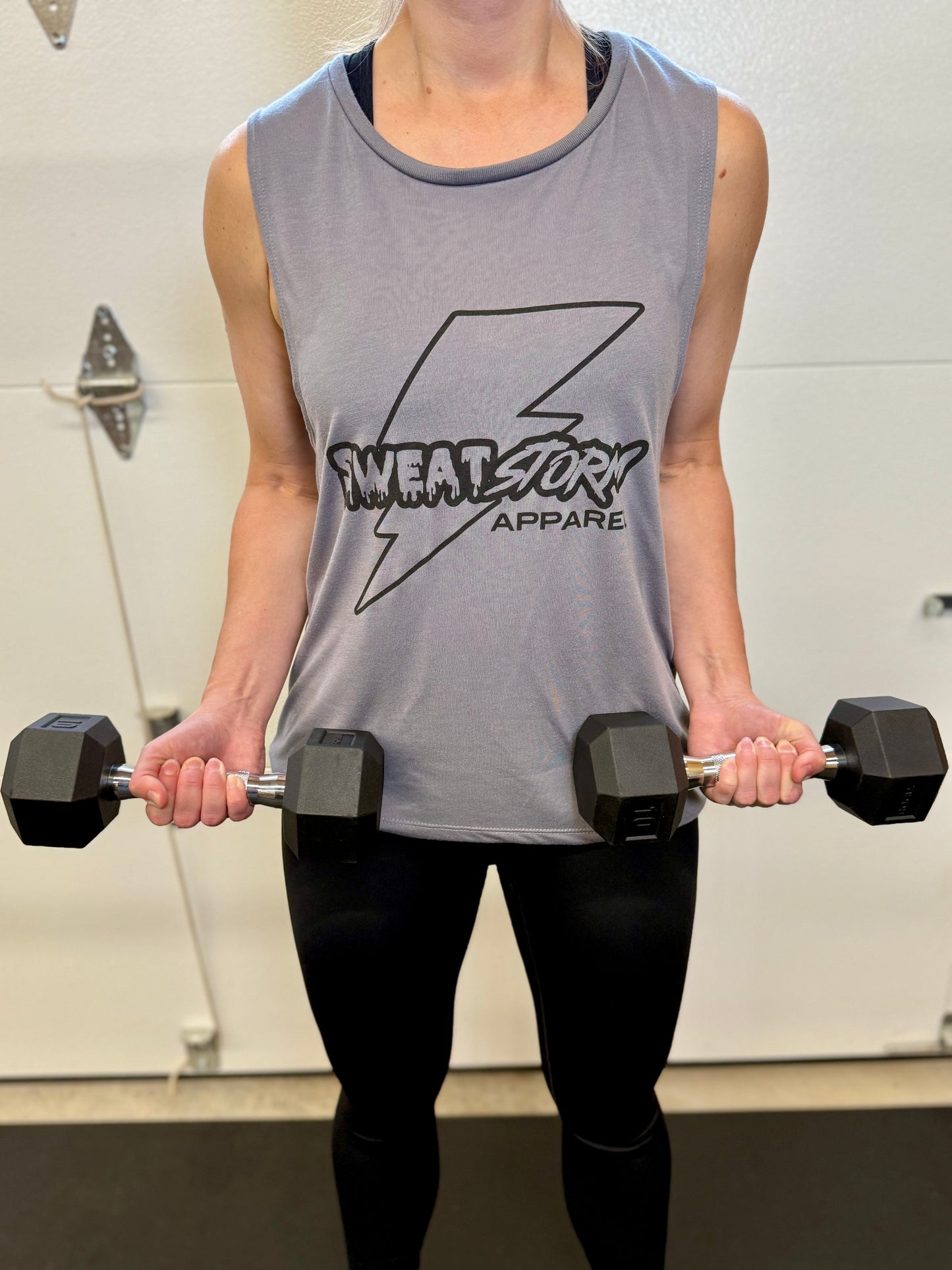 Person lifting dumbbells wearing a sleeveless shirt with branding in a gym setting