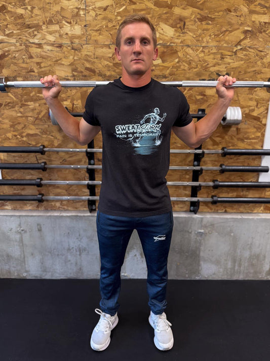 Man holding a barbell in a gym setting with wooden walls and weight racks wearing Sweat Storm Apparel blue joggers and black pain is temporary t-shirt