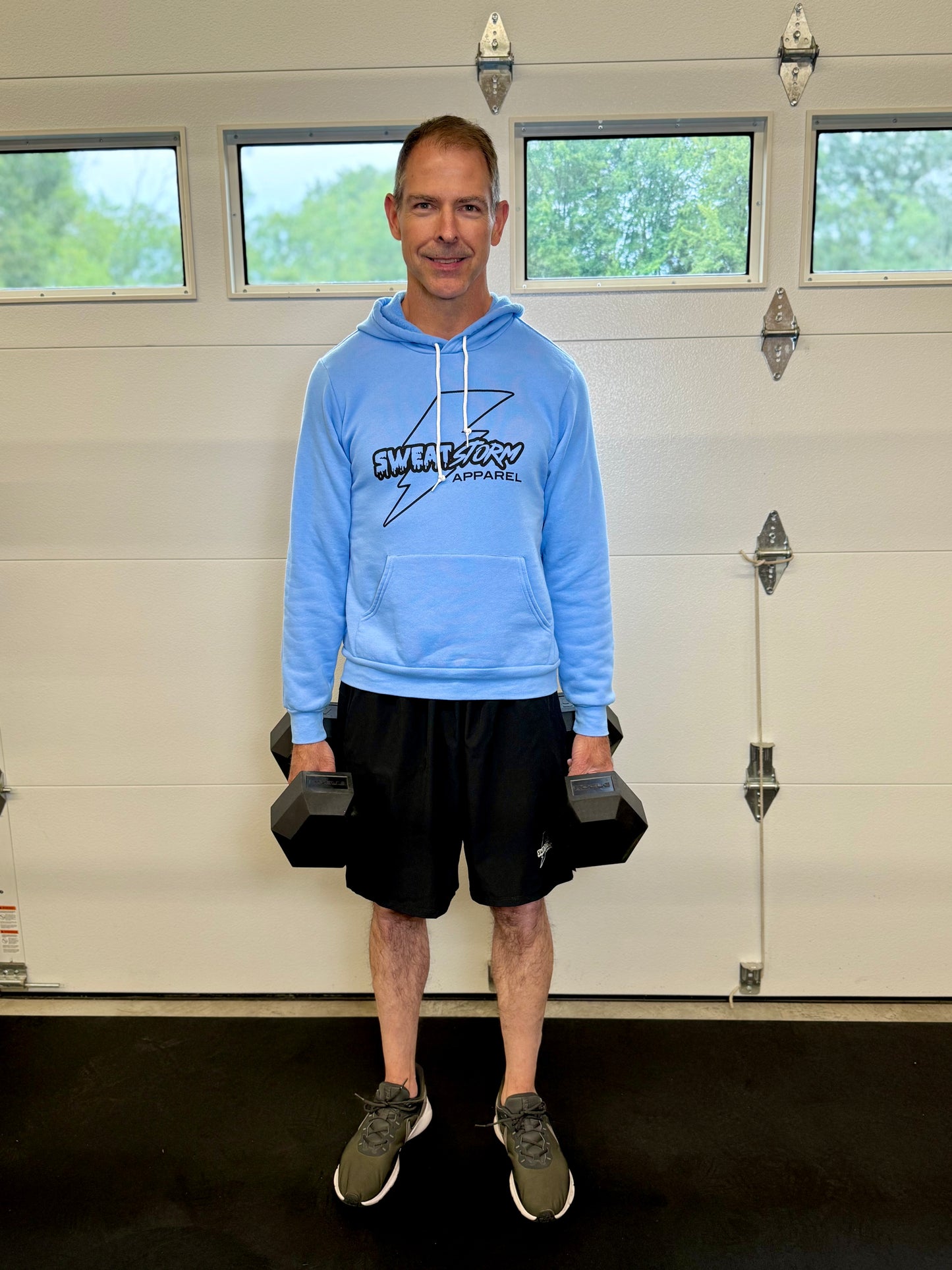 Man in blue Sweat Storm Apparel hoodie and black shorts holding dumbbells in front of a garage door.