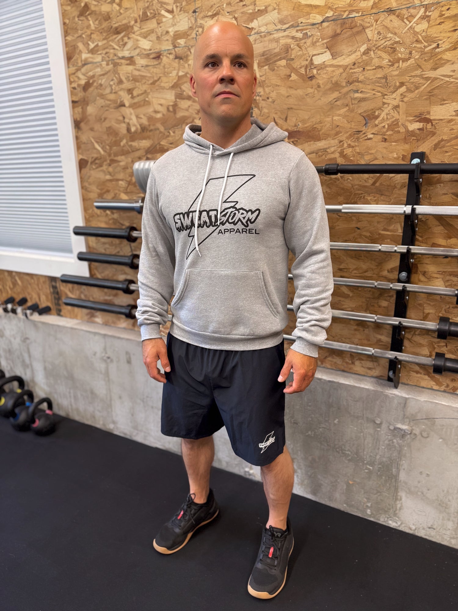 Man wearing a gray Sweat Storm Apparel hoodie with a logo in a gym setting