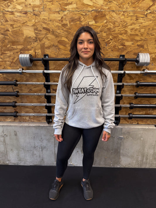 Person wearing a gray Sweat Storm Apparel hoodie with a logo in front of a wooden wall with barbells.
