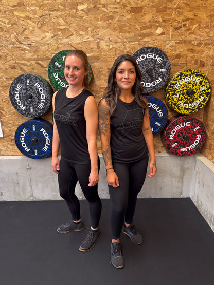 Two women standing in front of Rogue weight plates on a wooden wall in black Sweat Storm Apparel tank top