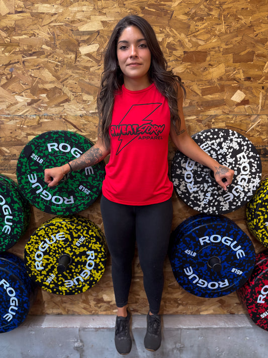 Woman in a red Sweat Storm Apparel logo tank top standing next to Rogue fitness weights against a wooden wall.