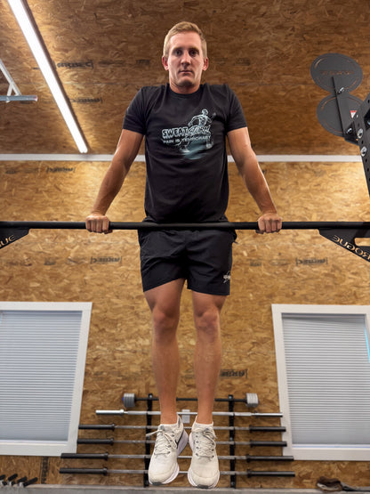 Man on a rogue rig in a gym setting with wooden walls and white windows wearing a black Sweat Storm Apparel t-shirt and shorts with Pain Is Temporary on the t-shirt