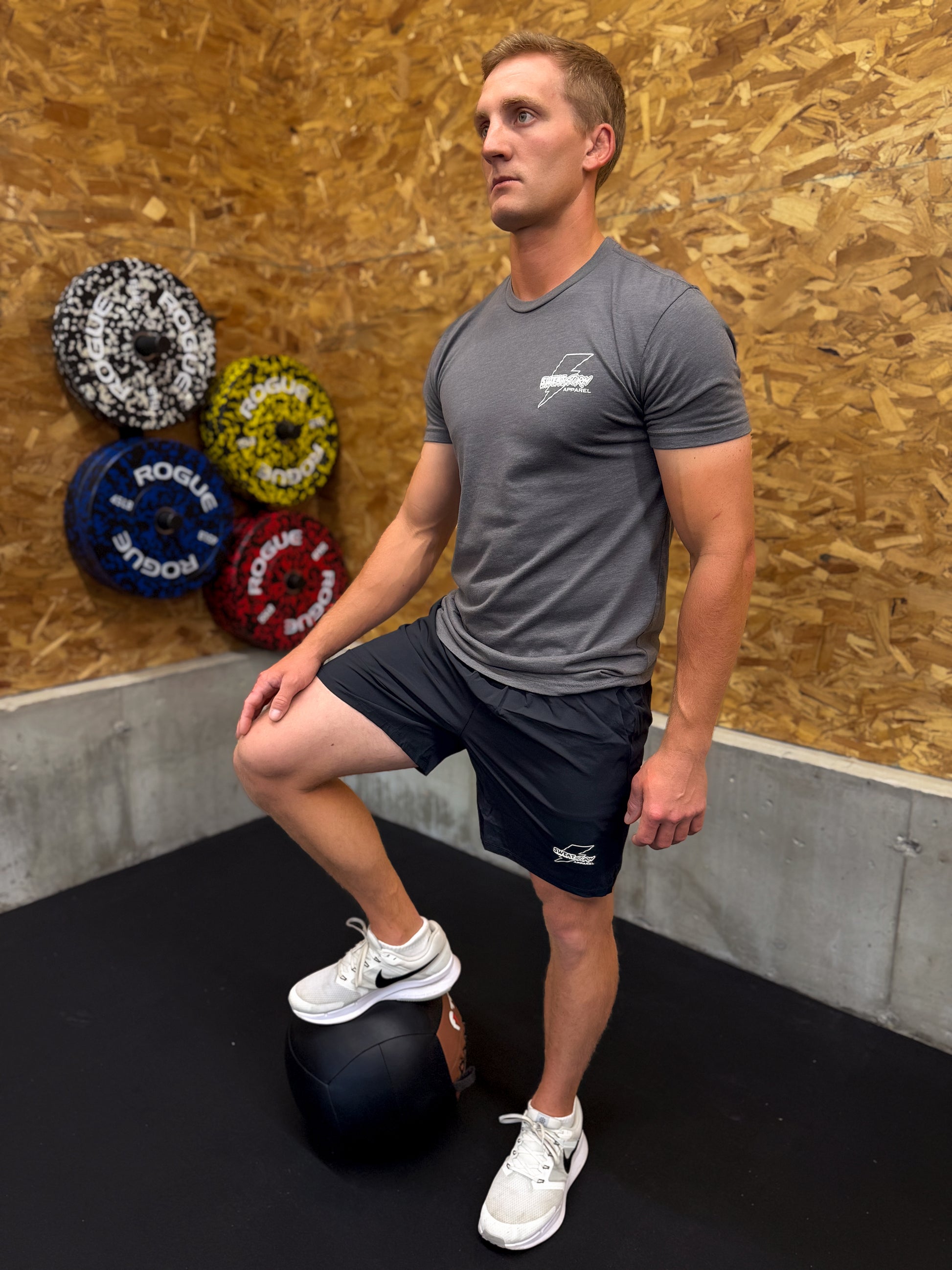 Man performing a leg exercise with a medicine ball in a gym setting wearing gray Sweat Storm Apparel t-shirt and black shorts