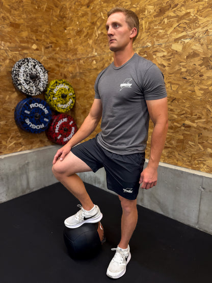 Man performing a leg exercise with a medicine ball in a gym setting wearing gray Sweat Storm Apparel t-shirt and black shorts