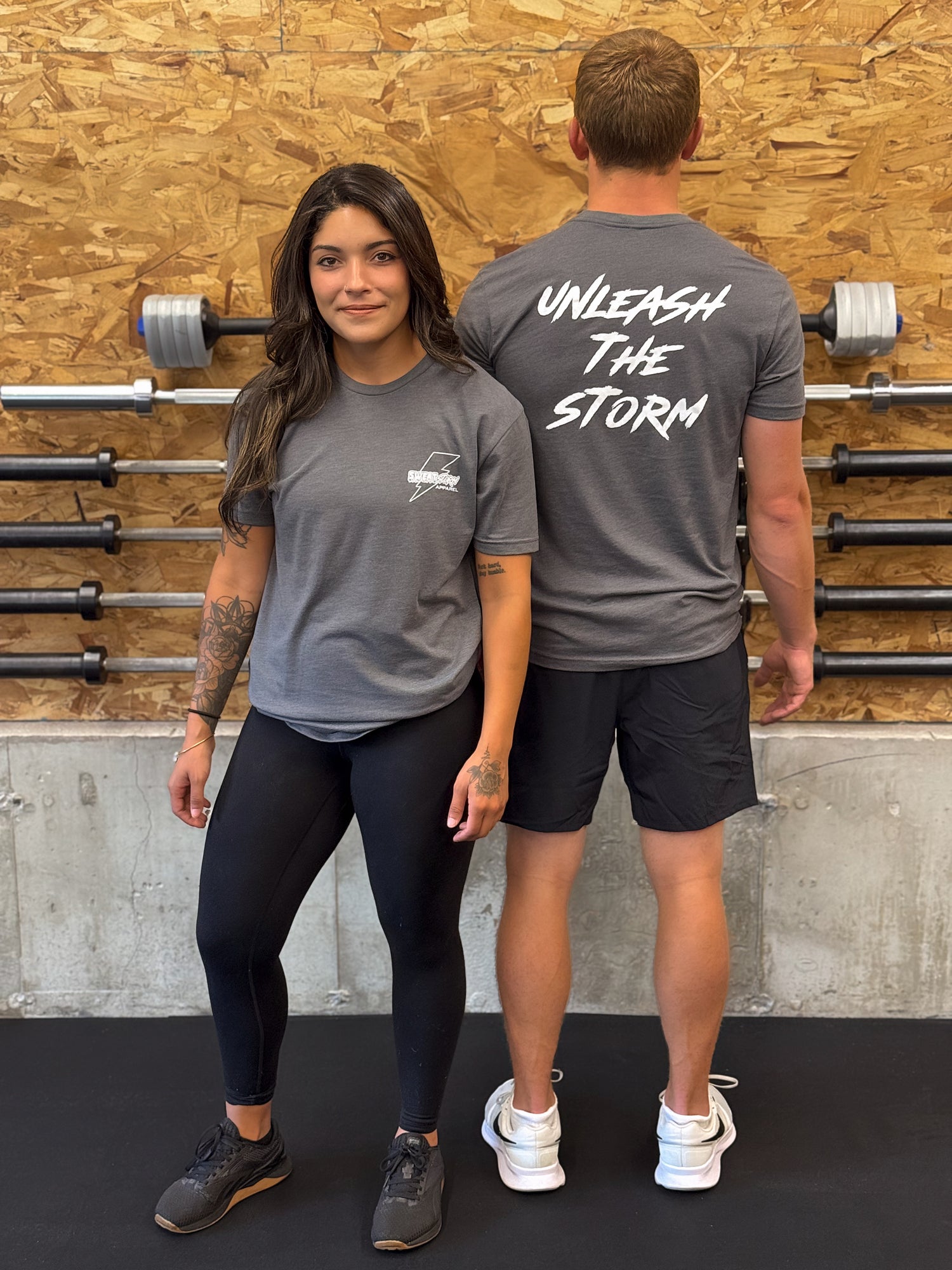 Two people wearing gray Sweat Storm Apparel unleash the storm t-shirts with text, standing in front of a wooden wall with gym equipment.