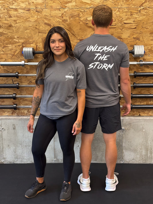 Two people wearing gray Sweat Storm Apparel unleash the storm t-shirts with text, standing in front of a wooden wall with gym equipment.