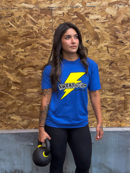 Person wearing a blue Sweat Storm Apparel logo t-shirt holding a kettlebell against a wooden panel background