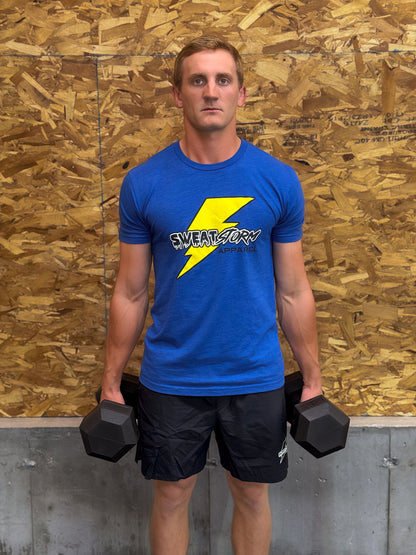 Man holding dumbbells wearing a blue Sweat Storm Apparel logo t-shirt against a wooden panel background