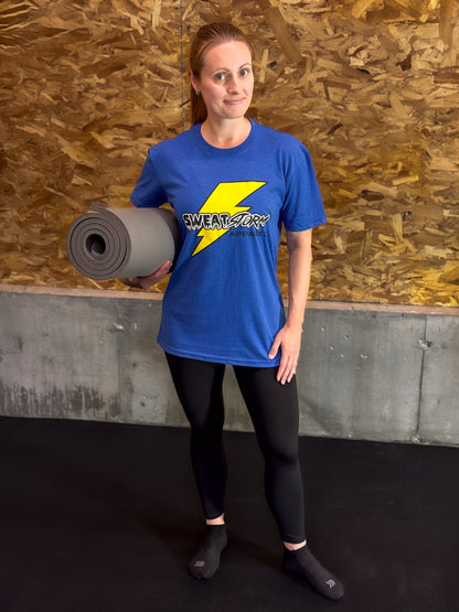 Person wearing a blue 'Sweat Storm Apparel' t-shirt holding a yoga mat against a wooden wall.
